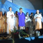 Starsza kobieta ubrana w granatową sukienkę przemawia do mikrofonu z młodą kobietą ubraną w biały kombinezon. Za nimi stoją młode dziewczyny ubrane na biało.