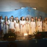 Grupa młodych ludzi ubranych na biało śpiewają i grają na scenie w sali szkolnej. Mężczyna w garniturze składa gratulacje.