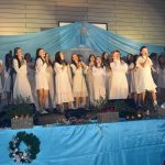 Grupa młodych ludzi ubranych na biało śpiewają i grają na scenie w sali szkolnej.