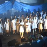 Grupa młodych ludzi ubranych na biało śpiewają i grają na scenie w sali szkolnej.