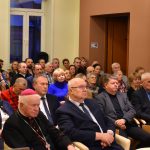 Grupa osób siedzi w jasnej sali na krzesełkach na konferencji.