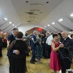 Grupa osób w eleganckich strojach, tańczących na rozległej sali.