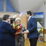 Mężczyzna w garniturze przekazuje kwiaty dwóm starszym paniom. W tle baner gminny oraz grupa osób zasiadająca na stołach.