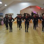 Grupa dziewcząt w strojach stylizowanych na kowbojskie tańcząca na parkiecie rozległej sali.