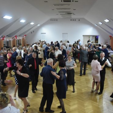 Grupa osób w eleganckich strojach, tańczących na rozległej sali.