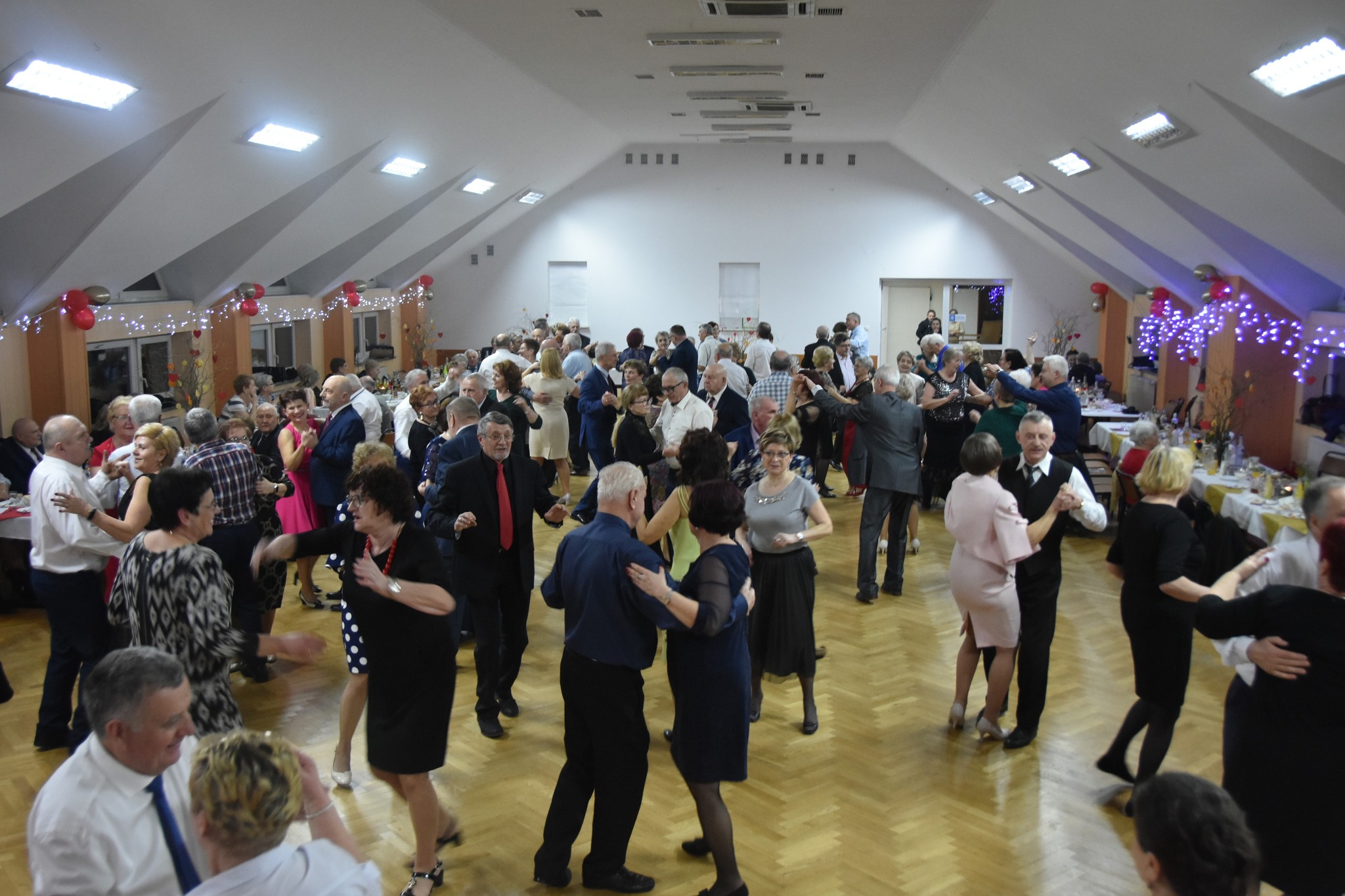 Grupa osób w eleganckich strojach, tańczących na rozległej sali.