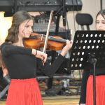 Grupa dziewcząt w eleganckich strojach podczas występu muzycznego. Jedna z nich gra na skrzypcach.