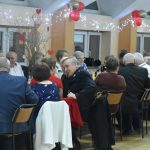 Grupa starszych osób obydwu płci zasiadająca przy podłużnych stołach bankietowych.