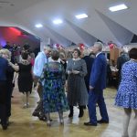 Grupa osób w eleganckich strojach, tańczących na rozległej sali.