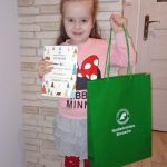 Dziewczynka o długich włosach ubrana w różową bluzę i spodniczkę szarą trzyma w rękach dyplom i zieloną torbę preznetową.