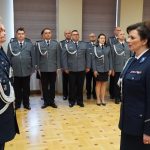 Ubrani w mundury policyjne mężczyzna i kobieta spoglądający na siebie. W tle grupa policjantów. Wszyscy stoją na sali konferencyjnej.