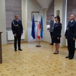 Mężczyzna w garniturze przemawia do mikrofonu. W te grupa sosób w mundurach policyjnych. Wszyscy znajdują się na sali konferencyjnej.