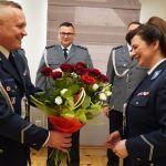 Męzczyzna w mundrze przekazuje podobnie ubranej kobiecie bukiet kwiatów. W tle dwójka mężczyzn w strojach policyjnych.