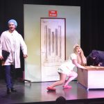 Młoda kobieta ubrana w strój pielęgniarki wraz z osoba w stroju grabarza przesuwają skrzynie ucharakteryzowaną na trumnę. Obok stoi młody mężczyzna w stroju lekarza. (aktorzy na scenie)