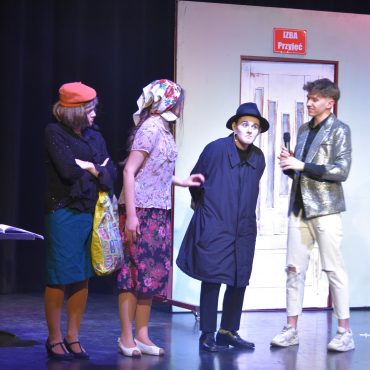 Dwie dziewczyny ubrane w stroje pań domu, osoba ubrana w długi, ciemny płaszcz i ciemna czapke oraz chłopak ubrany w srebrną marynarkę i białe wytarte jeansy (aktorzy na scenie).