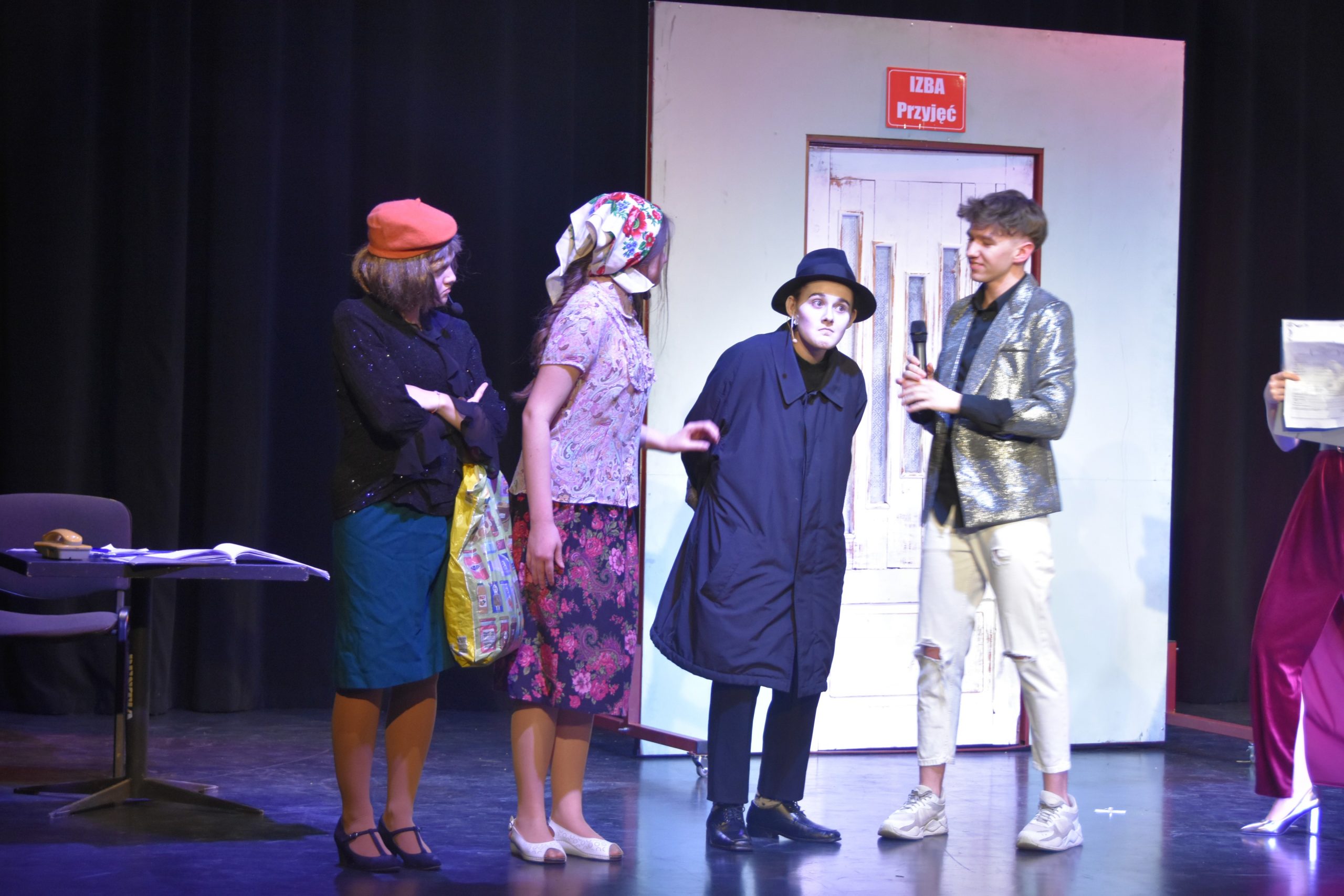 Dwie dziewczyny ubrane w stroje pań domu, osoba ubrana w długi, ciemny płaszcz i ciemna czapke oraz chłopak ubrany w srebrną marynarkę i białe wytarte jeansy (aktorzy na scenie).