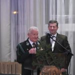 Dwóch mężczyzn w czarnych garniturach. Starszy przemawia za pomocą mikrofonu.