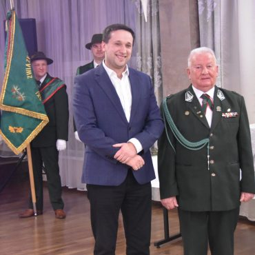 Dwóch mężczyzn w garniturach. W tle trzyosobowy poczet sztandarowy.