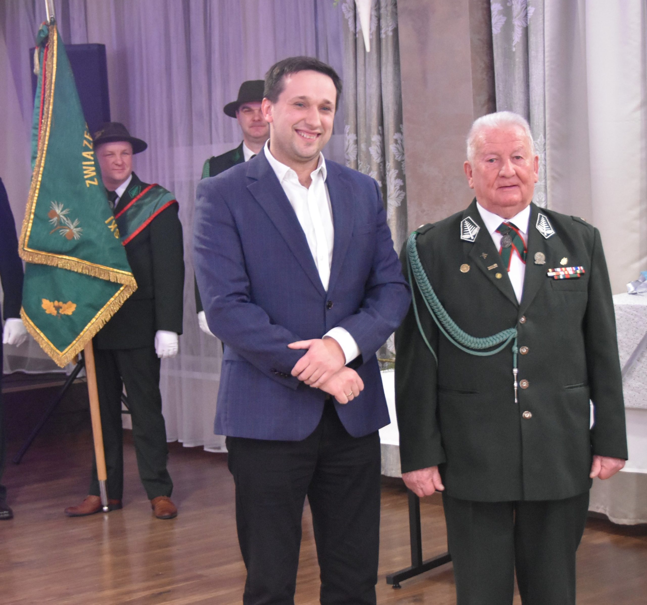 Dwóch mężczyzn w garniturach. W tle trzyosobowy poczet sztandarowy.