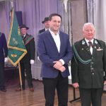 Dwóch mężczyzn w garniturach. W tle trzyosobowy poczet sztandarowy.