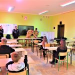 Grupa dzieci siedzących w ławkach w klasie. Z przodu stoi grupa elegancko ubranych dorosłych osób.