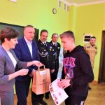 Mężczyzna w otoczeniu mezczyzn w graniturach i elegancko ubranej kobiety wręcza podarunek i dyplom młodemu chłopakowi w bluzie.