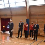 Grupa dorosłych osób w garniaturach i strojach sportowych na sali gimanstycznej.