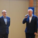 Dwóch dorosłych meżczyzn w garniturach. Jeden przemawia za pomocą mikrofonu.