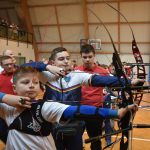 Grupa młodych łuczników strzelajaca z łuków sportowych na sali gimnastycznej.