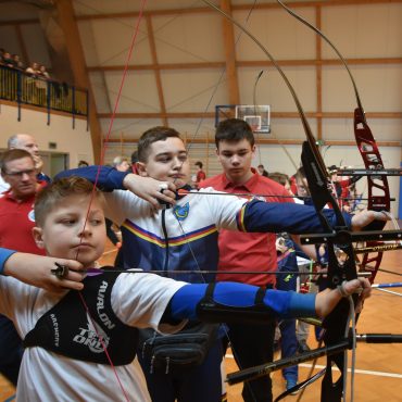 Grupa młodych łuczników strzelajaca z łuków sportowych na sali gimnastycznej.