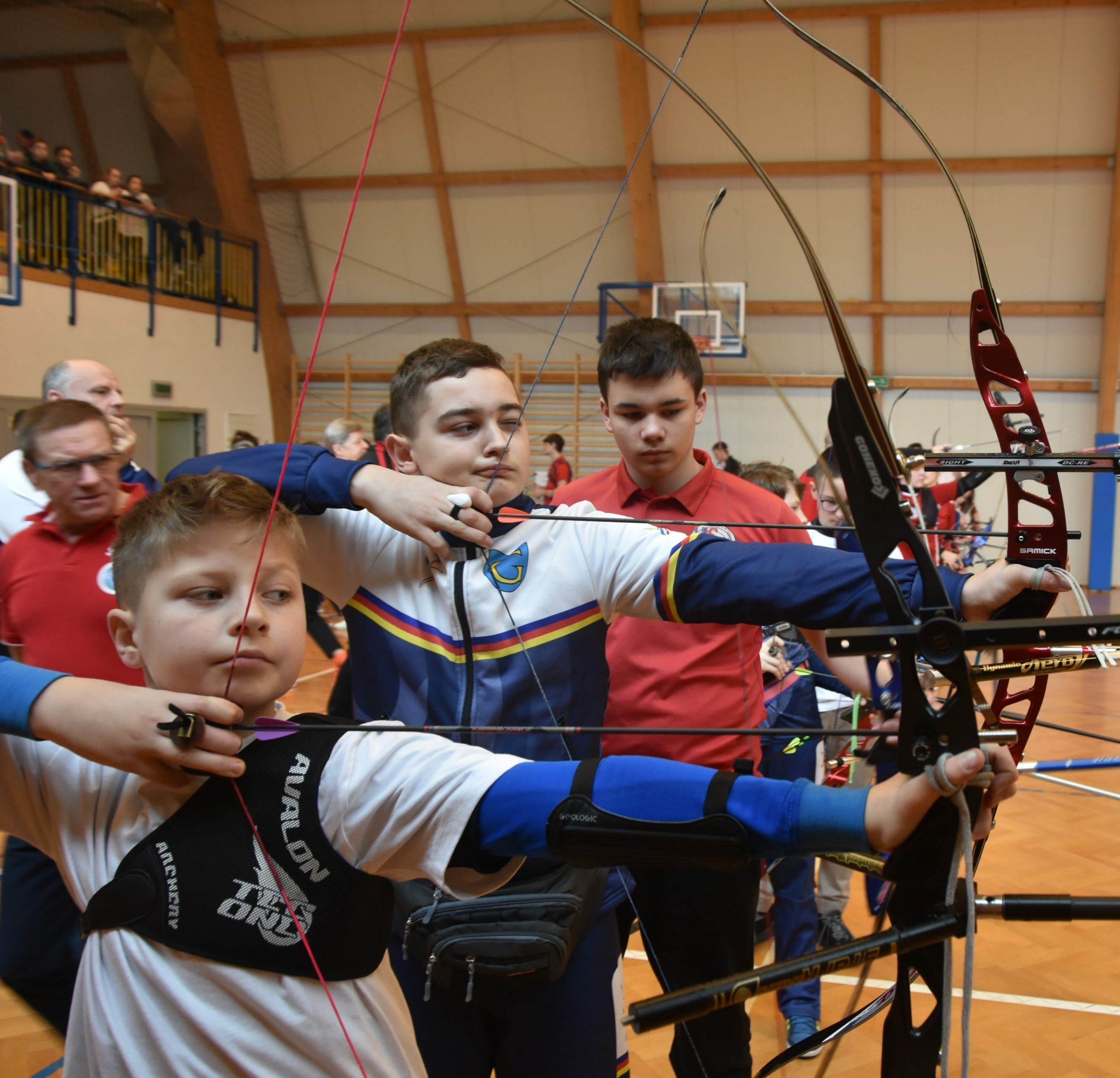 Grupa młodych łuczników strzelajaca z łuków sportowych na sali gimnastycznej.