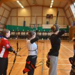 Grupa młodych łuczników strzela do tarczy w sali gimastycznej.