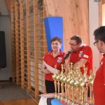 Dwoch mężczyzn i kobieta w strojach sędziowskich obok pucharów na sali gimastycznej.