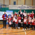 Grupa osób (głównie dzieci i młodzieży) w łuczniczych strojach sportowych na sali gimnastycznej.