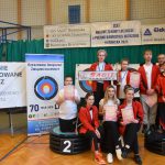 Grupa osób (głównie dzieci i młodzieży) w łuczniczych strojach sportowych na sali gimnastycznej.