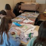 Grupa dzieci siedzi przy stolikach i wycina z papieru różne rzeczy.