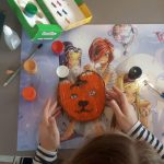 Na stole leżą różne przybory szkolne, Dziewczynka maluje na drewnie obrazek.