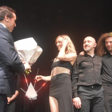 Męzczyzna w graniturze przekazuje bukiet kwiatów młodej wokalistce. Obok dóch mężczyzn w ciemnych strojach.