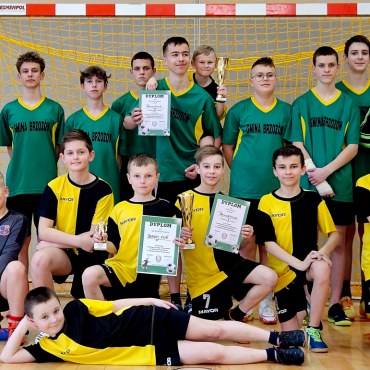 Grupa chłopców - piłkarzy pozujacych do zdjecia na sali gimnastycznej ze statuetką i dyplomami przed bramką. Obok stoi dorosły mężczyzna z długą czarną brodą.