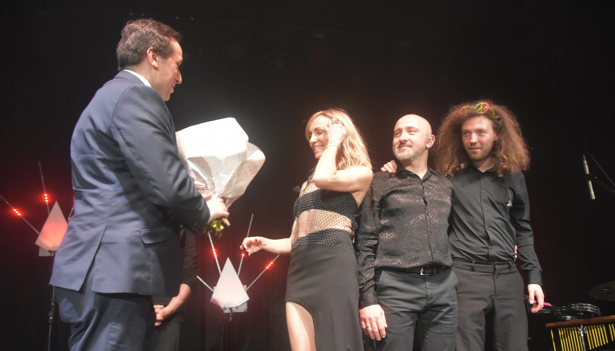 Męzczyzna w graniturze przekazuje bukiet kwiatów młodej wokalistce. Obok dóch mężczyzn w ciemnych strojach.