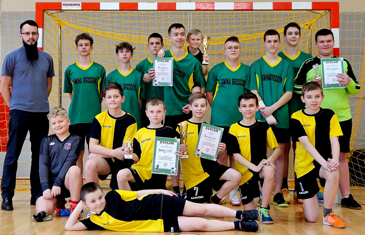 Grupa chłopców - piłkarzy pozujacych do zdjecia na sali gimnastycznej ze statuetką i dyplomami przed bramką. Obok stoi dorosły mężczyzna z długą czarną brodą.