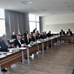 Grupa dorosłych osób w eleganckich ubraniach na sali konferencyjnej. Na stole znajdują się dokumenty i butelki wody.