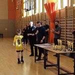 Grupa dorosłych na sali gimanstycznej oraz stolik z pucharami i dyplomami. Młody zawodnik podchodzi do meżczyzny z mikrofonem by odebrać nagrodę.