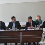 Grupa dorosłych osób w eleganckich ubraniach na sali konferencyjnej. Na stole znajdują się dokumenty i butelki wody.