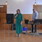 Kobieta w zielonej sukni przemaiwa za pomoca mikrofonu na rozległej sali. Z tyłu stoi męzczyzna w jasnej koszuli.