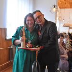 Kobieta w zielonej koszuli i mężczyzna w graniturze pozujący do zdjęcia. Kobieta trzyma aniołka i słodycze.