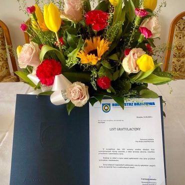 List gratulacyjny i bukiet kwaitów położone na stole.