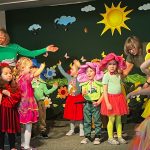 Grupa dzieci w stroju kwiatów i owadów na scence teatralnej na sali, wraz z opiekunami.