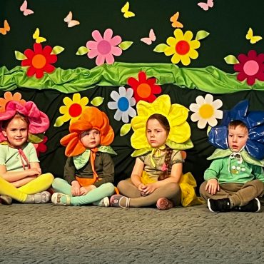 Kilkoro dzieci w stroju kwaitów siedzaca na scenie.