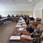 Grupa dorosłych osób w eleganckich ubraniach na sali konferencyjnej. Na stole znajdują się dokumenty i butelki wody.